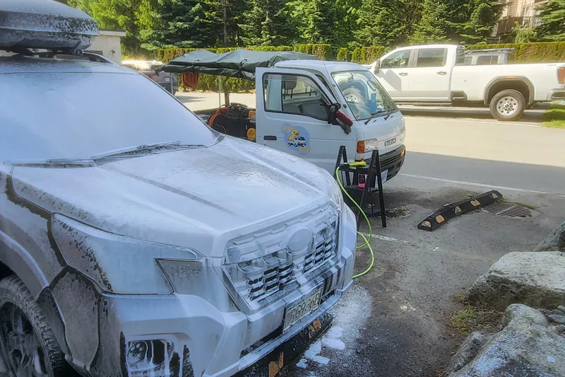 A roof-boxed SUV under foam on a residential street, Duty Duck kei truck set up behind it.